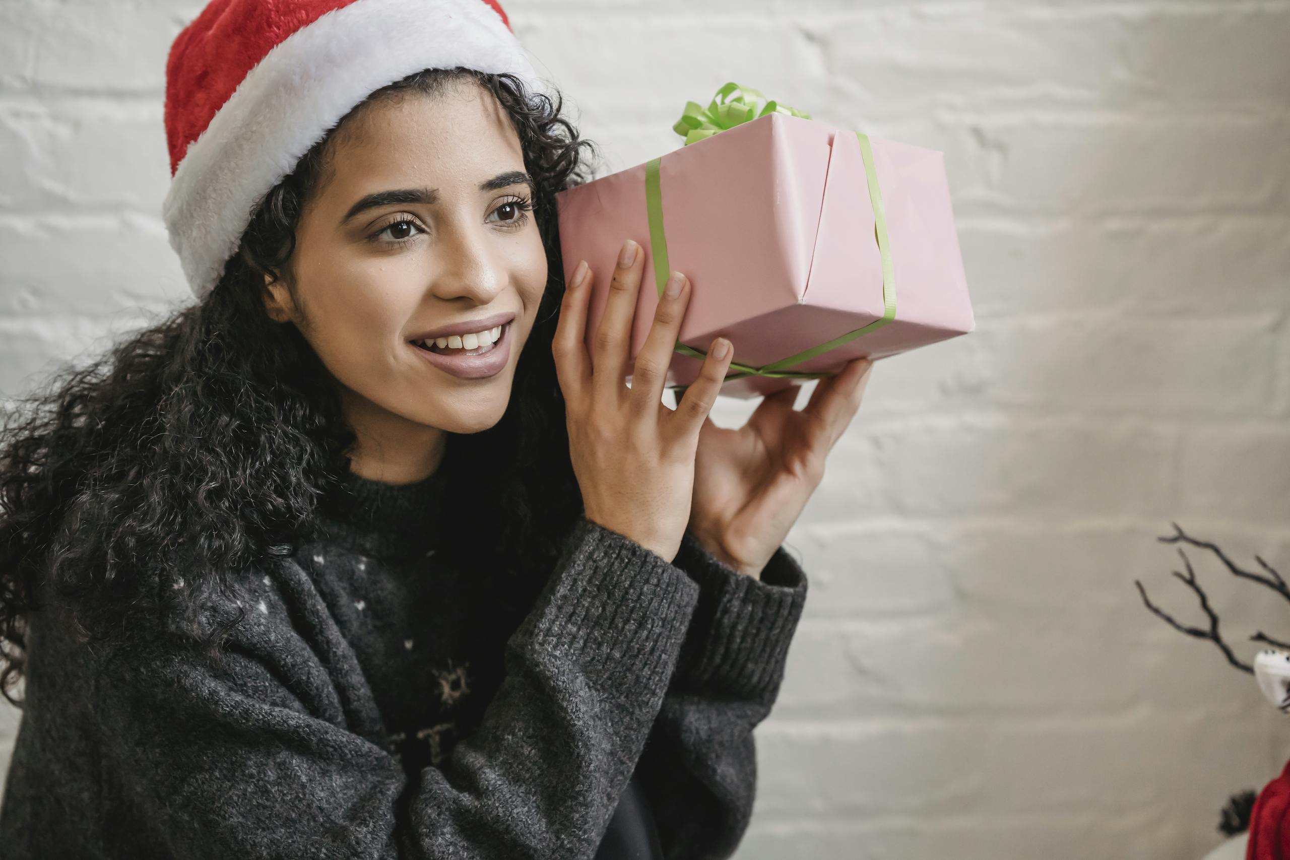 Smiling young ethnic female with curly hair wearing Santa hat and holding wrapped gift box near ear while looking away in excitement during Christmas party